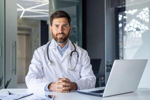 retrato do uma sorridente confiante masculino médico dentro uma branco casaco sentado às uma escrivaninha dentro uma clínica e olhando às a Câmera. foto