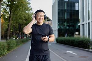 retrato do uma sorridente jovem ásia homem corrida em uma cidade rua vestindo fones de ouvido e uma telefone, ouvindo para música on-line. foto