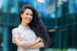 retrato do feliz e bem sucedido o negócio mulher, patrão dentro camisa sorridente e olhando às Câmera dentro escritório com cruzado braços, hispânico mulher com encaracolado cabelo ao ar livre. foto