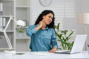 uma profissional mulher às uma escrivaninha caretas com pescoço dor enquanto trabalhando em uma computador portátil dentro a escritório contexto. foto