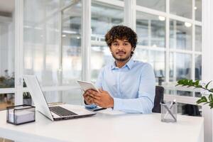 uma focado jovem homem dentro uma azul camisa acionado dentro multitarefa com uma computador portátil e tábua às uma brilhante escritório escrivaninha. foto