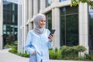 uma jovem lindo o negócio mulher dentro uma hijab anda em através a cidade, uma muçulmano mulher detém uma telefone dentro dela mãos, usa a inscrição em uma Smartphone, sorrisos, navega a Internet. foto