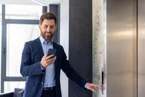 uma sorridente jovem masculino homem de negocios é em pé dentro a escritório espaço, chamando a elevador e usando uma Móvel telefone. foto