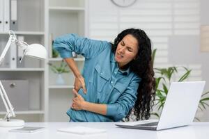 uma mulher dentro uma azul camisa sente forte costas dor, estremecendo dentro desconforto enquanto sentado às dela escritório escrivaninha com uma computador portátil. foto