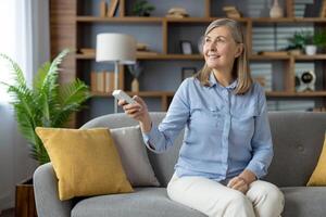 feliz idosos mulher sentado em uma sofá, usando uma controlo remoto ao controle para ajustar a ar condicionador dentro uma acolhedor vivo quarto contexto. foto