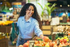 feliz mulher comprador dentro supermercado, latim americano mulher compra maçãs, frutas e vegetais, coloca dentro a ecológico saco foto