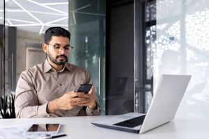 sério concentrado homem de negocios às a local de trabalho dentro a escritório, a homem detém uma Smartphone dentro dele mãos, uma trabalhador com uma computador portátil navega a Internet e social redes, tipos uma texto mensagem. foto