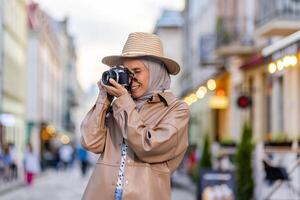 jovem lindo mulher caminhando dentro a tarde cidade dentro hijab, turista com Câmera e vestindo uma chapéu inspeciona a histórico cidade sorridente com satisfação, muçulmano mulher em uma viagem. foto