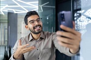 jovem bem sucedido homem de negocios às local de trabalho dentro escritório falando em ligar usando aplicativo em telefone, fechar acima homem às local de trabalho sorridente. foto