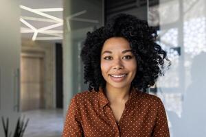 retrato do uma confiante jovem africano americano mulher com encaracolado cabelo, sorridente calorosamente dentro uma moderno escritório contexto. ela desgasta uma à moda polca ponto camisa e exala profissionalismo e acessibilidade. foto