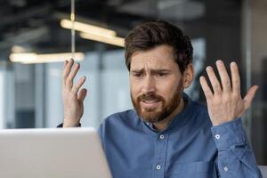 uma confiante jovem homem senta dentro uma moderno escritório e parece desapontadamente às a computador portátil tela, espalhando dele mãos. fechar-se foto. foto