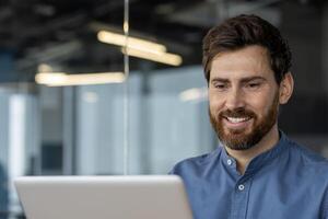 uma profissional masculino dentro uma inteligente azul camisa às a escritório, sorridente enquanto olhando às dele computador portátil, exalando confiança e positividade. foto