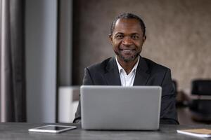 uma homem dentro uma terno é sentado às uma escrivaninha com uma computador portátil e tábua. ele é sorridente e ele é feliz foto