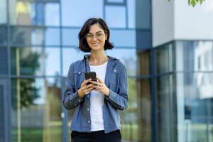 confiante jovem empresária com óculos usando uma Smartphone lado de fora uma moderno escritório prédio. isto imagem captura uma casual ainda profissional vibe. foto