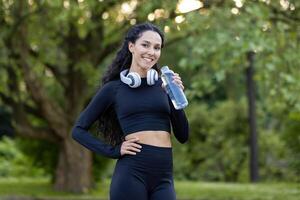 alegre ginástica entusiasta leva uma descansar dentro uma verde parque, segurando uma água garrafa com fones de ouvido por aí dela pescoço. foto