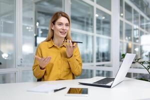 sorridente jovem o negócio mulher trabalhando dentro a escritório em uma computador portátil, sentado às a mesa e falando em a telefone através a Alto-falante. foto