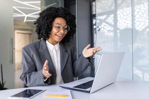 sorridente jovem afro-americano o negócio mulher sentado dentro a escritório às a escrivaninha e falando em uma conexão, conduzindo a conectados conferência. foto