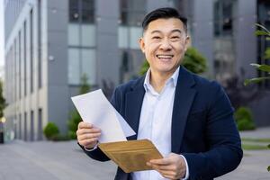 retrato do sorridente jovem homem de negocios homem em pé lado de fora escritório, segurando documentos e aberto envelope, recebido Boa notícias, parece alegremente às Câmera. foto