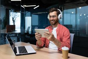 bem sucedido homem de negocios às local de trabalho sorridente homem assistindo conectados sentado às escrivaninha vestindo fones de ouvido e óculos programador segurando tábua computador dentro escritório. foto