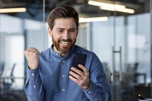 feliz e sorridente jovem homem sentado dentro escritório moderno Centro e olhando às telefone, desfrutando sucesso e mostrando vitória gesto com mão. fechar-se foto. foto