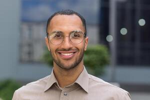 fechar-se foto retrato do jovem empreendedor vestindo copos, hispânico homem sorridente e olhando às Câmera, comece empreendedor lado de fora moderno escritório construção