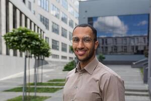 fechar-se foto retrato do jovem empreendedor vestindo copos, hispânico homem sorridente e olhando às Câmera, comece empreendedor lado de fora moderno escritório construção