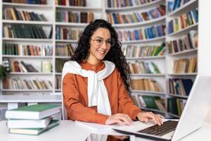 lindo latim americano aluna estudando dentro universidade biblioteca, mulher com encaracolado cabelo e óculos digitando em computador portátil teclado sorridente, tendo Diversão preparando para exame. foto