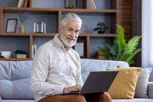 a idosos homem com uma caloroso sorrir sentado em uma sofá, usando uma computador portátil dentro uma elegantemente mobiliado vivo sala, cercado de pacífico casa decoração. foto