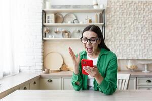 lindo jovem mulher dentro óculos e verde camisa ler Boa notícia a partir de vermelho telefone foto