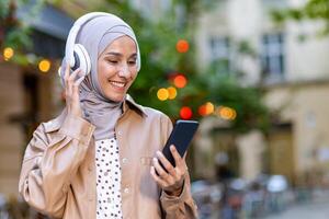 jovem lindo mulher caminhando dentro a cidade, muçulmano mulher dentro hijab em tarde andar dentro fones de ouvido ouvindo para música sorridente alegremente usando Smartphone aplicativo. foto