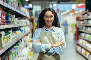 retrato do hispânico mulher vendedor dentro supermercado, fêmea trabalhador dentro avental com encaracolado cabelo e óculos sorridente e olhando às Câmera, vendedora entre bens com família produtos químicos e prateleiras foto