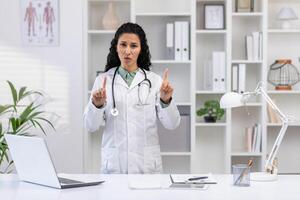 retrato do fêmea médico dentro clínica escritório, mulher infeliz e chateado olhando às Câmera apontando dedos gesto não, trabalhando às escrivaninha com computador portátil. foto