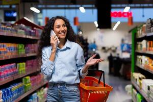 sorridente hispânico mulher comprador dentro supermercado, anda em entre linhas do prateleiras com bens, mulher alegremente fala em Móvel telefone, comprador recomenda loja para amigo foto