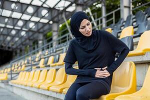 uma fêmea atleta vestindo uma hijab experiências desconforto e estômago dor enquanto sentado sozinho dentro uma estádio com esvaziar assentos. foto