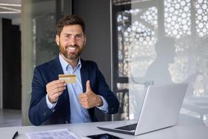 retrato do uma bem sucedido e sorridente jovem homem homem de negocios, banco empregado sentado às uma escrivaninha dentro a escritório, segurando uma dourado crédito cartão e mostrando uma super com dele dedo para a Câmera. foto