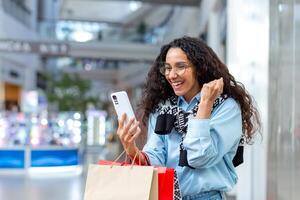 retrato do feliz mulher comprador, hispânico mulher dentro shopping center, usa Smartphone, navega conectados descontos e vendas, detém mão acima, comemora recebido oferecer, ganhar foto