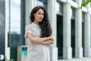 retrato do uma confiante empresária com cruzado braços em pé lado de fora do uma contemporâneo escritório prédio, exibindo profissionalismo e força. foto