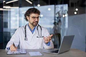 profissional masculino médico com estetoscópio envolve dentro uma ligar consulta com uma paciente usando dele computador portátil dentro uma moderno clínica escritório. foto