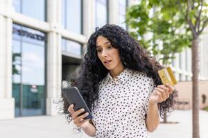 chateado e desapontado mulher recebido dinheiro transferir erro, hispânico mulher com banco crédito cartão e telefone enganado caminhando baixa rua lado de fora escritório prédio. foto