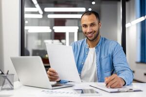 concentrado indiano homem analisando documentos enquanto trabalhando em dele computador portátil dentro uma moderno vivo quarto configurar. foto