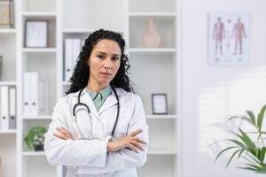 fechar-se retrato do sério e confiante hispânico fêmea doutor, mulher olhando às Câmera com cruzado braços pensativo, trabalhando às local de trabalho dentro clínica escritório, consultando pacientes. foto