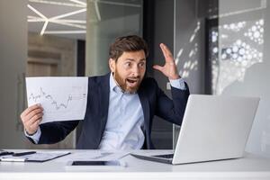 uma profissional homem parece Bravo e exasperado durante uma ligar reunião, segurando financeiro gráficos e gesticulando com dele mão. foto