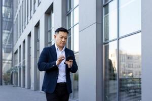 uma jovem ásia homem dentro uma o negócio terno é caminhando baixa a rua perto escritório edifícios e usando uma Móvel telefone. foto