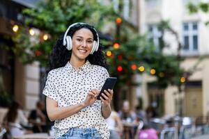 jovem latim americano empresária com encaracolado cabelo, desfrutando música com fones de ouvido, usando Smartphone dentro a cidade. foto