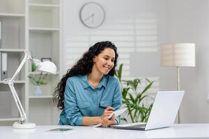 uma jovem mulher sorrisos enquanto assistindo uma webinar em dela computador portátil e levando notas, sentado às uma bem organizado casa escritório escrivaninha. foto