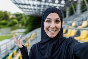 uma alegre muçulmano mulher dentro uma hijab captura uma selfie momento com uma Largo sorrir às uma ensolarado Esportes estádio com amarelo assentos. foto