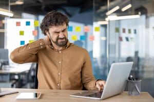 uma masculino escritório empregado sente afiado pescoço dor enquanto trabalhando em dele computador portátil, indicando a precisar para ergonômico espaços de trabalho. foto