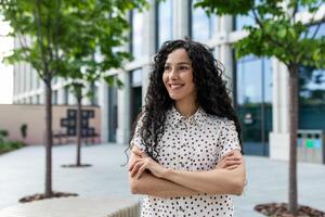 jovem feliz hispânico fêmea aluna com braços cruzado lado de fora universidade campus sorridente contente e olhando ausente. foto