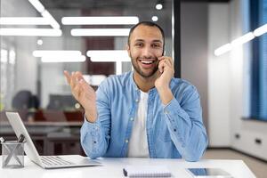 feliz masculino profissional dentro uma casual camisa tendo uma agradável conversação em Móvel telefone enquanto trabalhando dentro uma brilhante contemporâneo escritório. foto