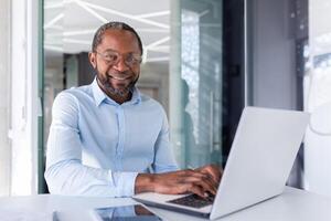 bem sucedido africano americano homem de negocios dentro camisa e óculos trabalhando dentro brilhante moderno escritório, homem sorridente e olhando às Câmera, retrato do bem sucedido patrão usando computador portátil sentado às ambiente de trabalho. foto